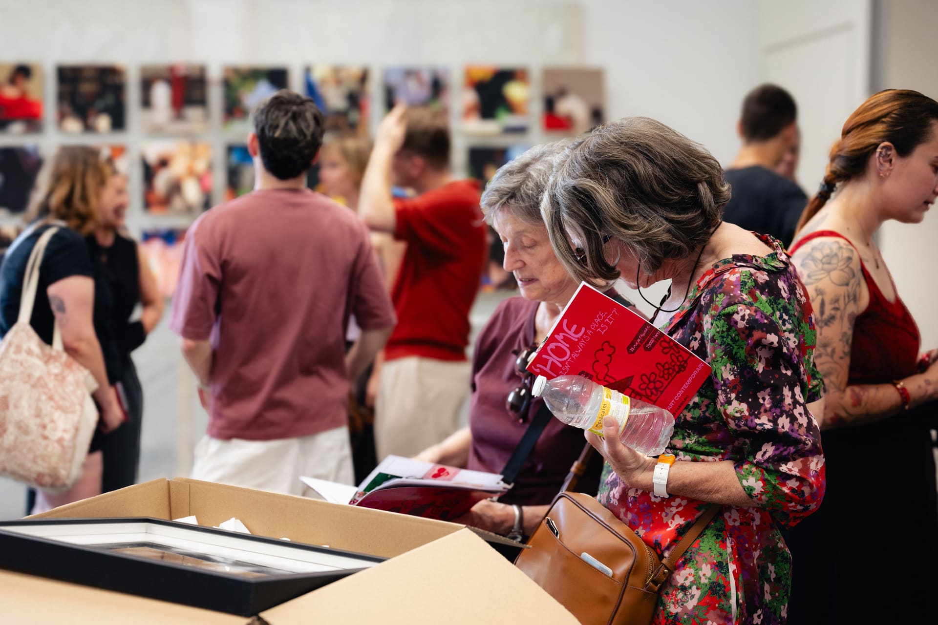 Visiteuses feuilletant le livret rouge de l’exposition devant le mur de tirages, au Kiosque Citoyen.